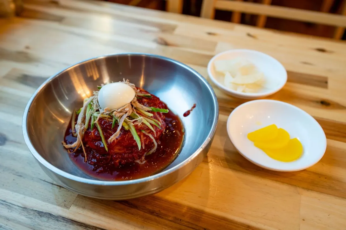 金属碗中的辣拌釜山小麦冷面，上有鸡蛋、黄瓜和肉丝