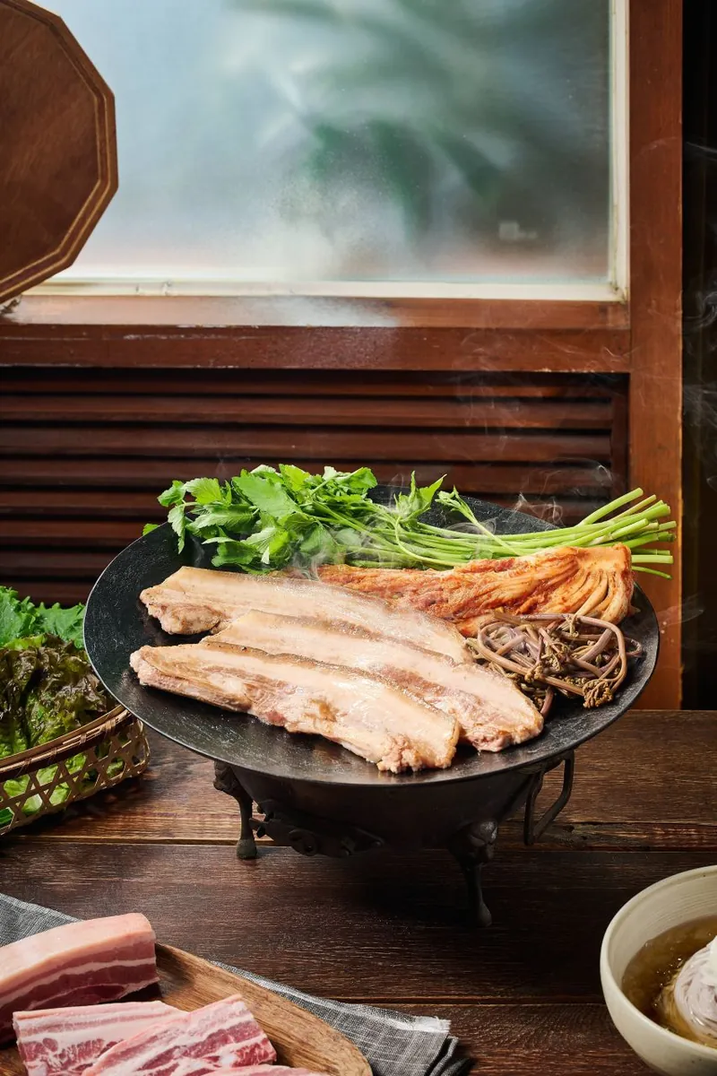 Các dải thịt ba chỉ nướng trên vỉ tại bàn, cạnh rau và món phụ