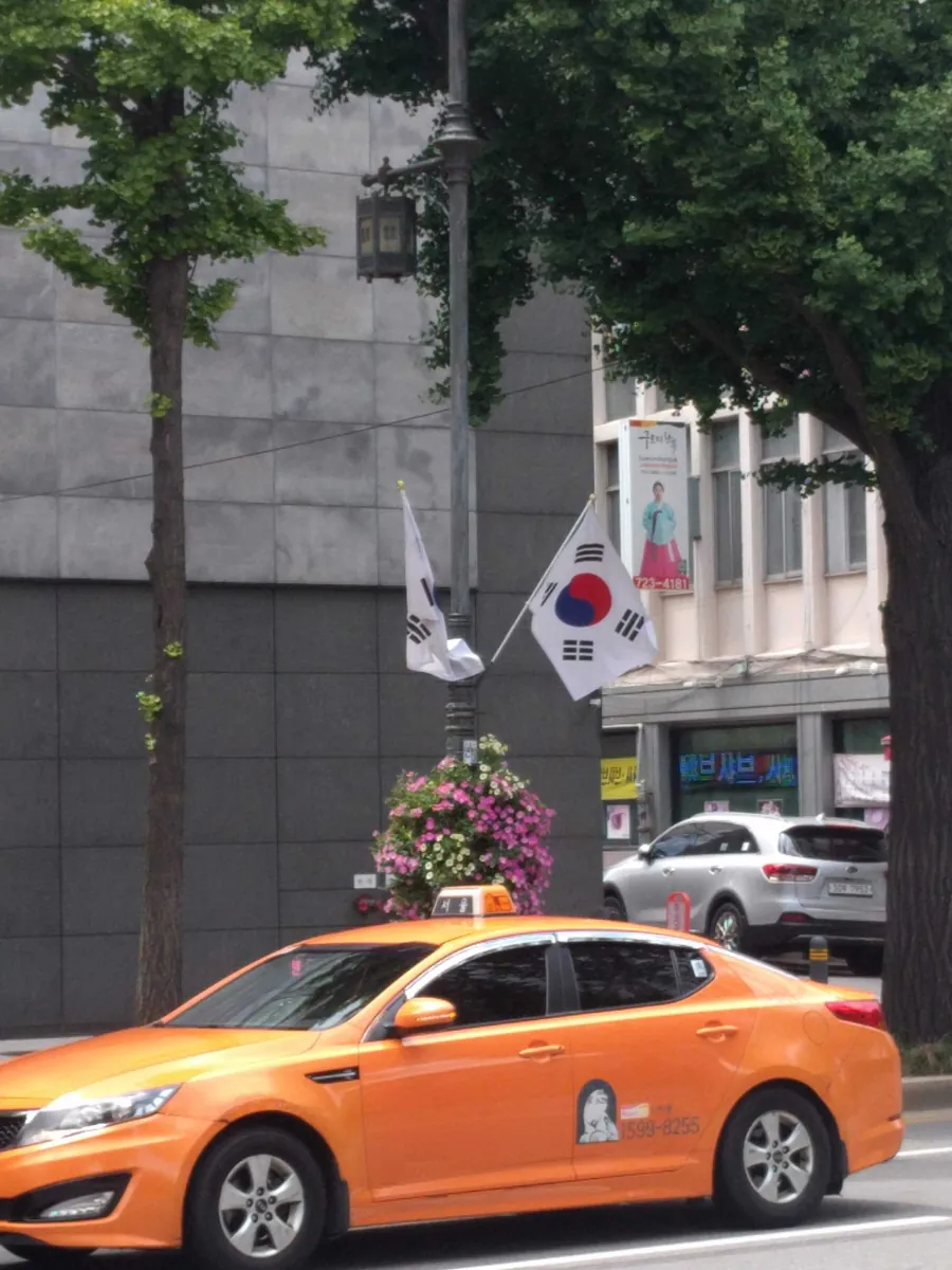 Taxi màu cam ở Seoul đi qua tòa nhà đá cạnh các lá cờ Hàn Quốc